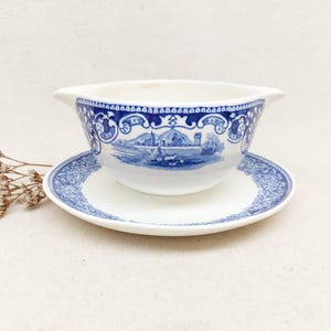 May include: A white and blue porcelain gravy boat with a matching saucer. The gravy boat features a blue and white floral design and a scene of a town with a church and houses. The saucer has a blue and white floral design.