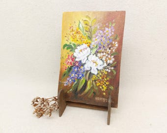May include: A small floral oil painting on canvas, presented on a wooden easel. The artwork depicts a bouquet of white, yellow, and purple flowers with green foliage. The artist's signature, "VANSUIEL", is visible on the bottom right of the painting.
