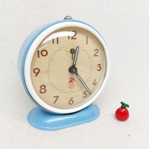 May include: A vintage blue alarm clock with white trim and a cream-coloured face. The clock has black numbers and hands, and a small red logo with the letters "STD".