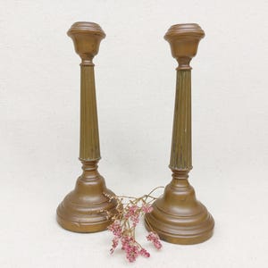 Può includere: Due portacandele in metallo dorato con colonne scanalate e basi rotonde. I portacandele sono su una superficie bianca.