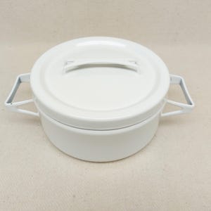 Op de afbeelding: Een witte emaille kookpot met bijpassende deksel. De pot heeft twee handvatten aan de zijkant en een handvat op het deksel. De pot is rond en lijkt leeg te zijn. De pot staat op een neutraal gekleurd oppervlak.