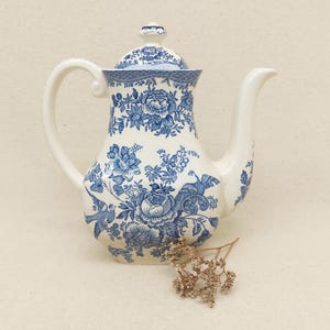 Può includere: Una teiera in ceramica bianca con un motivo floreale blu e bianco. La teiera ha un coperchio e un beccuccio. La teiera è appoggiata su una superficie bianca.