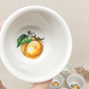 Pode incluir: Uma tigela de cerâmica branca com um desenho pintado de um pêssego com folhas verdes. A tigela está vazia e tem um acabamento liso e brilhante.
