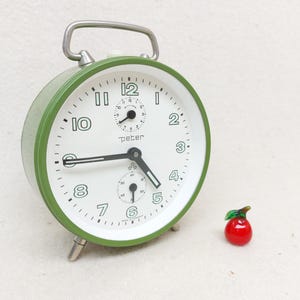 Könnte beinhalten: Ein grüner Wecker mit weißem Zifferblatt und schwarzen Zeigern. Die Uhr hat eine Sekundenzeiger und ein kleines Zifferblatt zum Einstellen des Alarms. Die Uhr steht auf einer weißen Oberfläche und daneben liegt ein roter Glasapfel.