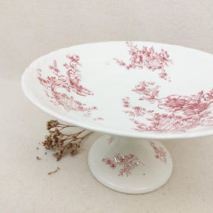 Op de afbeelding: Witte keramische taartstandaard met een ronde voet en een brede, geschulpte rand. De standaard is versierd met een rood bloemmotief. De taartstandaard is een decoratief stuk voor het presenteren van desserts of andere items.