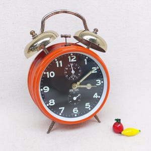May include: A retro orange alarm clock with a black face and white numerals. The clock features gold-coloured bells and a handle. A small red and yellow glass fruit decoration is next to the clock.