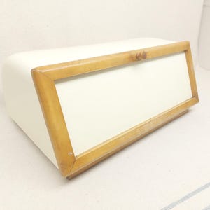 Op de afbeelding: Crèmekleurige broodtrommel met een houten frame rond het voorpaneel. De trommel heeft een gebogen bovenkant en een rechthoekige vorm. Het houten frame is lichtbruin van kleur. De broodtrommel staat op een wit oppervlak.