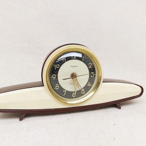 May include: A vintage Rhythm clock with a brown and cream-colored base. The clock face is black with gold trim and white numerals. The clock has a gold-colored second hand and hour and minute hands. The clock is approximately 8 inches wide.