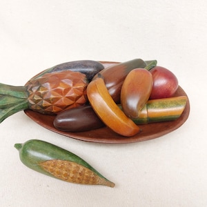 May include: A wooden bowl filled with hand-carved wooden fruit and vegetables. The bowl contains a pineapple, banana, eggplant, peppers, and an apple. The fruit and vegetables are painted in various shades of brown, green, red, and yellow.