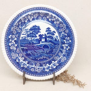 Può includere: Un piatto in porcellana blu e bianco con un bordo floreale decorativo e un'immagine centrale di un ponte su un ruscello con alberi e un piccolo edificio.