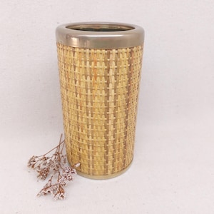 Op de afbeelding: Een cilindervormige vaas met een geweven rotan buitenkant en een metalen rand. De vaas is ongeveer 25 cm hoog en 13 cm in diameter. Een klein takje gedroogde bloemen rust bij de voet van de vaas. Woondecoratie.