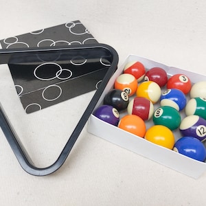 Könnte beinhalten: Ein Satz Billardkugeln in einer weißen Box, mit einem schwarzen Dreiecksgestell und einer schwarz-weiß gemusterten Box. Die Kugeln sind nummeriert und farbig, darunter rot, gelb, blau, grün und weiß.