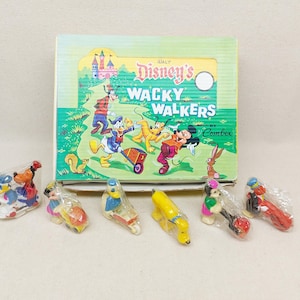 Könnte beinhalten: Ein Vintage-Spielzeugset von Disney's Wacky Walkers Comex. Die Box zeigt eine farbenfrohe Illustration von Disney-Figuren. Mehrere einzeln verpackte Plastikspielzeuge befinden sich vor der Box, darunter Donald Duck, Mickey Mouse und Pluto.