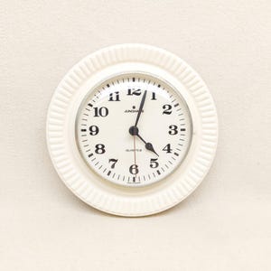 May include: A round wall clock with a cream-colored, ribbed frame and a white face. The clock features black Arabic numerals from 1 to 12, black hour and minute hands, a thin red second hand, and the text Junghans and Quartz printed in black.