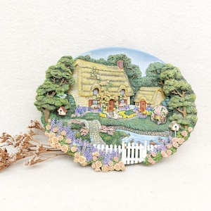 Pode incluir: Cena decorativa de uma casa de campo com telhado de colmo, árvores, flores e um riacho. O relevo detalhado apresenta uma cerca branca, casas de pássaros e um céu azul. Um raminho de flores secas em primeiro plano.