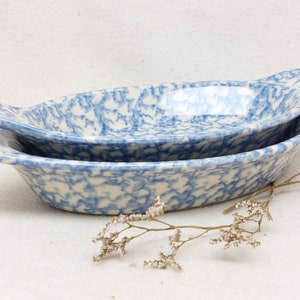 Op de afbeelding: Twee ovale keramische ovenschaaltjes met handgrepen, voorzien van een blauw-wit gespikkeld sponsmotief. De schaaltjes zijn gestapeld, waarbij de kleinere in de grotere rust, met een glanzende afwerking en gebogen randen.