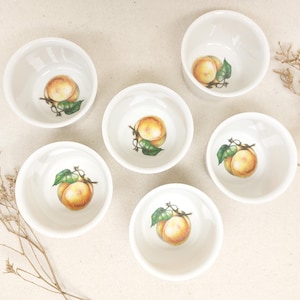 May include: Six white ceramic ramekins with a peach design. Each ramekin features a peach with green leaves and a stem.