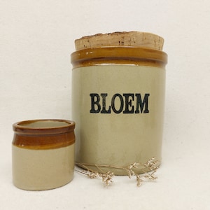 May include: A large beige ceramic canister with a brown rim and the word BLOEM printed in black, topped with a cork lid, accompanied by a smaller matching beige and brown ceramic pot and a small dried sprig of white flowers.