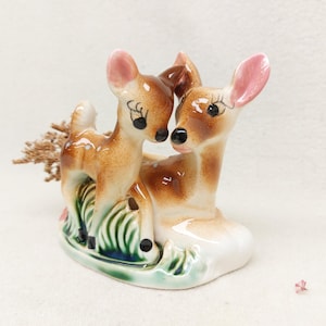 Puede incluir: Figura de cerámica de dos ciervos. El ciervo más grande está tumbado y el más pequeño está de pie junto a él. Los ciervos son marrones con manchas blancas y orejas rosas, sobre una base verde y blanca.