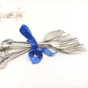 Pode incluir: Uma coleção de garfos prateados amarrados com uma vibrante fita de veludo azul. As alças dos garfos apresentam desenhos decorativos de conchas e flores. Os dentes dos garfos são visíveis, sugerindo seu uso para servir ou comer.