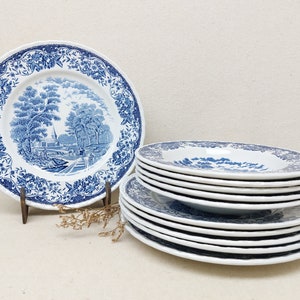 Puede incluir: Una pila de platos de cena de cerámica con estampado azul y blanco. Los platos tienen un diseño floral azul y blanco con una escena de un pueblo en el centro.