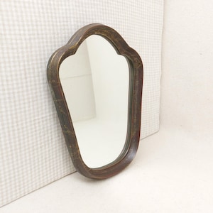 Pode incluir: Um espelho de parede com uma moldura de madeira castanho-escura com uma silhueta assimétrica de forma distinta. A moldura apresenta sinais visíveis de desgaste e uma textura envelhecida, rodeando uma superfície de vidro clara e refletora.