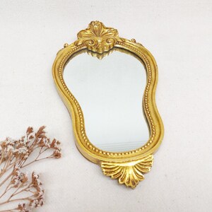 Peut inclure: Un petit miroir doré orné, de forme ondulée unique. Le cadre décoratif et détaillé présente des motifs floraux en haut et en bas. Le miroir est posé sur un fond blanc.