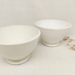 Może przedstawiać: Dwie kremowobiałe ceramiczne miski z podstawą na cokole. Miski mają zaokrąglony kształt i lekko teksturowaną powierzchnię. Gałązka suszonych kwiatów umieszczona jest w pobliżu misek.