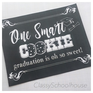 May include: A black chalkboard sign with white text that reads "One Smart Cookie graduation is oh so sweet!"  The sign features a drawing of a cookie with chocolate chips and a graduation cap.