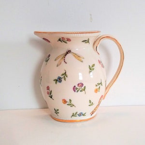 May include: A white ceramic jug with a floral and dragonfly design. The jug has a gold rim and a handle.