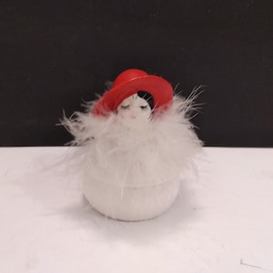 May include: A white ceramic figurine of a woman wearing a red hat and a white feather boa. The figurine is round and has a closed-eye expression.