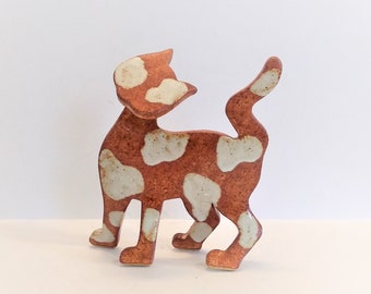May include: A ceramic cat figurine with a white and brown spotted pattern. The cat is standing on all four legs with its tail curled up.