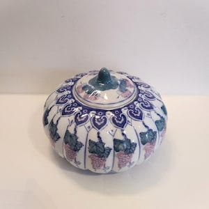 Peut inclure: Un pot en céramique blanche avec un couvercle, décoré de vignes de raisin bleues et violettes et d'un motif de feuilles vertes. Le pot a une forme ronde, semblable à une citrouille.