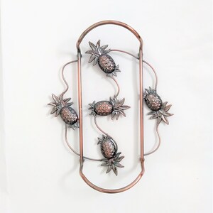 May include: A copper-toned metal pineapple-shaped wine bottle holder with five pineapples. The holder is designed to hold two bottles of wine.