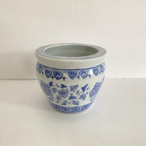 May include: A white ceramic planter with a rounded body and a wide opening. The exterior features blue hand-painted floral patterns, a geometric border around the rim, and a repeating rectangular motif along the base of the vessel.