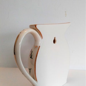 May include: White ceramic jug with a gold trim and floral design. The jug has a hole in the back for hanging on a wall.