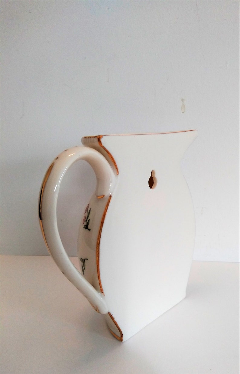 May include: White ceramic jug with a gold trim and floral design. The jug has a hole in the back for hanging on a wall.