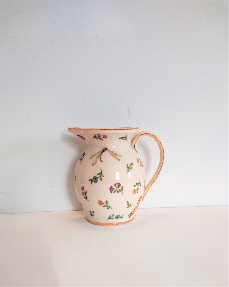 May include: A white ceramic jug with a floral and dragonfly design. The jug has a gold rim and a handle.