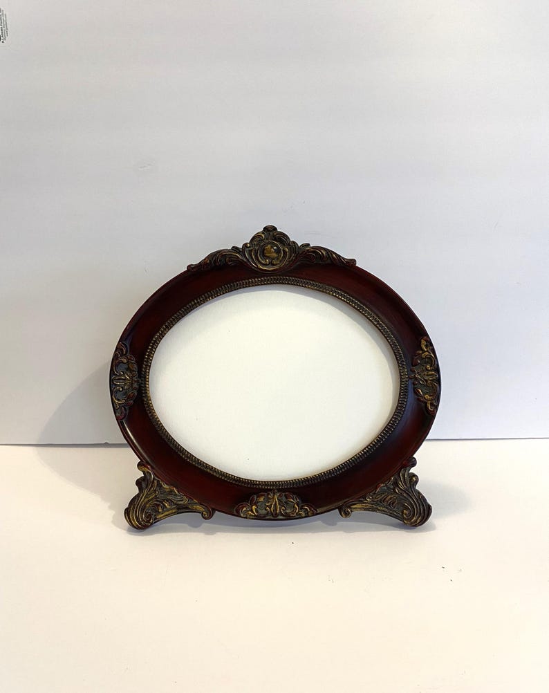 May include: An oval-shaped picture frame with a dark wood finish and ornate gold accents. The frame has a stand and is ready to display a photo or artwork.