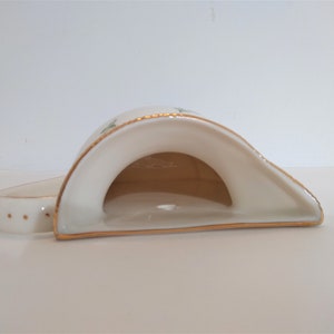May include: A white ceramic napkin holder with a gold trim and a floral design. The holder is shaped like a crescent moon and has a small opening in the centre.