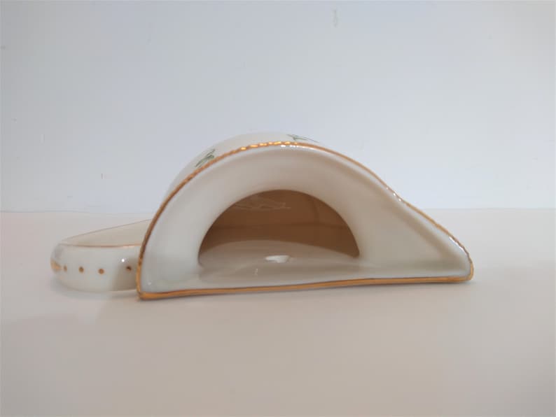May include: A white ceramic napkin holder with a gold trim and a floral design. The holder is shaped like a crescent moon and has a small opening in the centre.