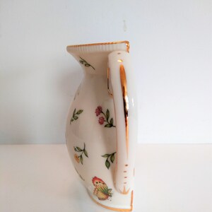May include: A white ceramic jug with a gold trim and a floral design. The jug has a handle with a gold trim and a butterfly design.