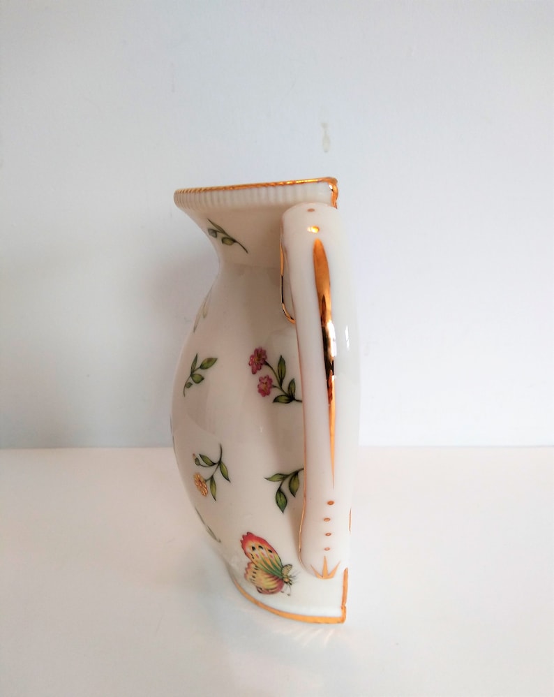 May include: A white ceramic jug with a gold trim and a floral design. The jug has a handle with a gold trim and a butterfly design.
