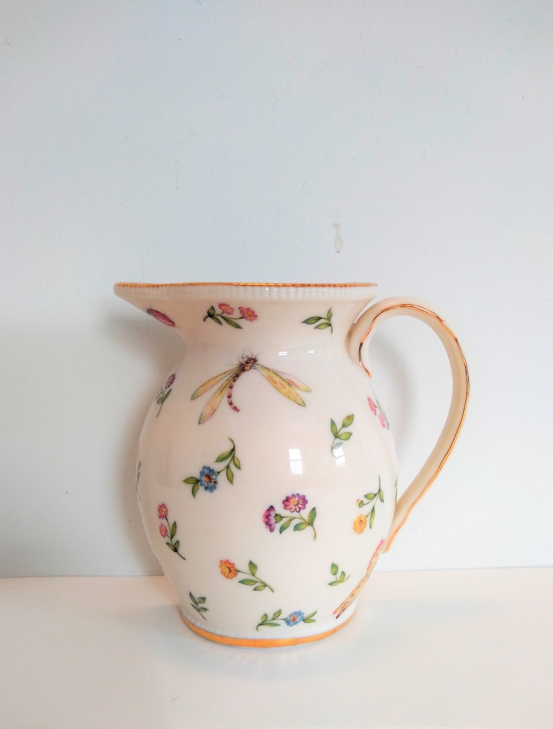 May include: A white ceramic jug with a gold rim and handle. The jug is decorated with a floral pattern and a dragonfly.