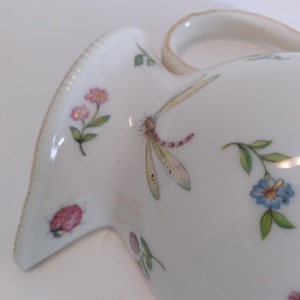 May include: White ceramic jug with a floral design featuring pink, blue, and yellow flowers, a dragonfly, and a ladybug. The jug has a gold-trimmed handle.