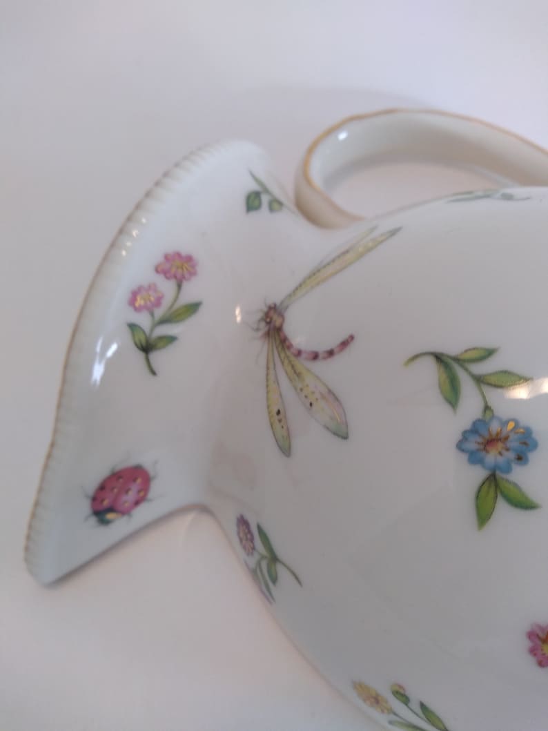 May include: White ceramic jug with a floral design featuring pink, blue, and yellow flowers, a dragonfly, and a ladybug. The jug has a gold-trimmed handle.