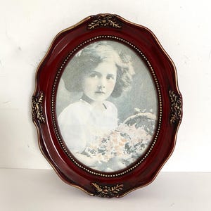 May include: A burgundy-coloured oval picture frame featuring a vintage photograph of a young girl holding flowers. The frame has ornate gold accents and a scalloped edge, creating a classic, antique aesthetic.