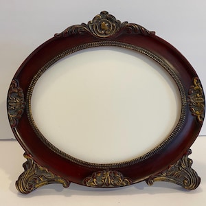 May include: An oval-shaped picture frame with a dark red finish and ornate gold accents. The frame has a stand and is made of wood.