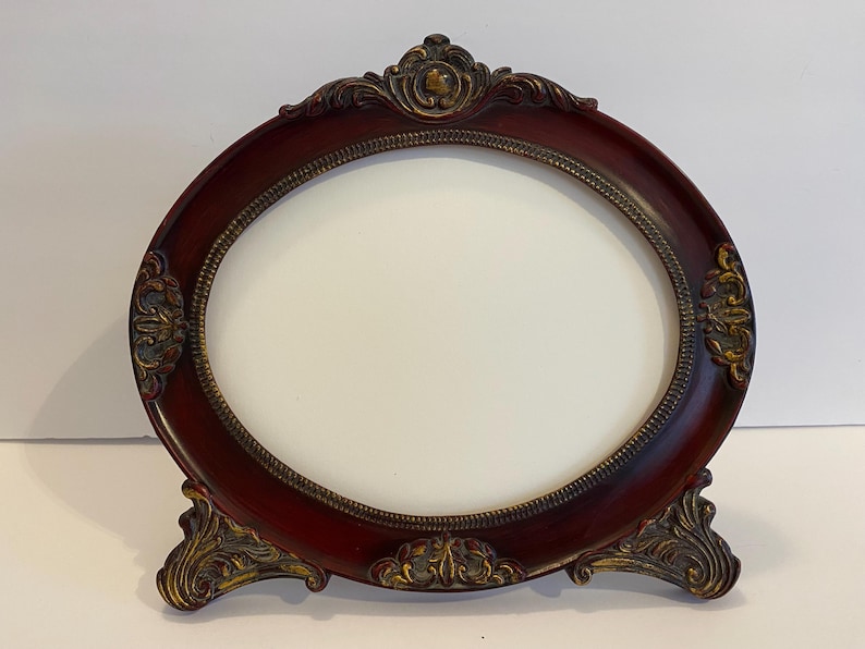 May include: An oval-shaped picture frame with a dark red finish and ornate gold accents. The frame has a stand and is made of wood.
