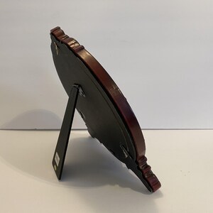 May include: A dark brown wood picture frame with a black backing and a decorative ornate design. The frame has a stand on the back to prop it up.